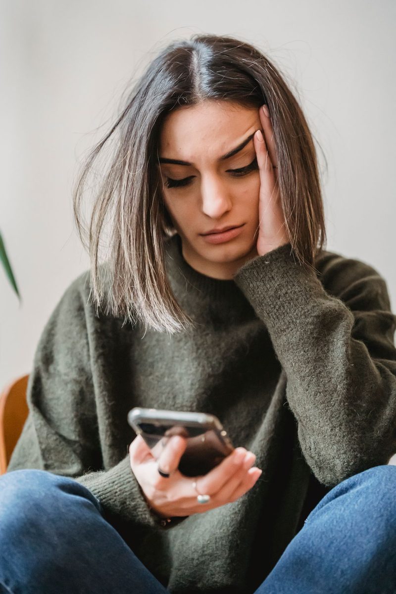Young woman sitting indoors with a hand on her head, frowning at her smartphone