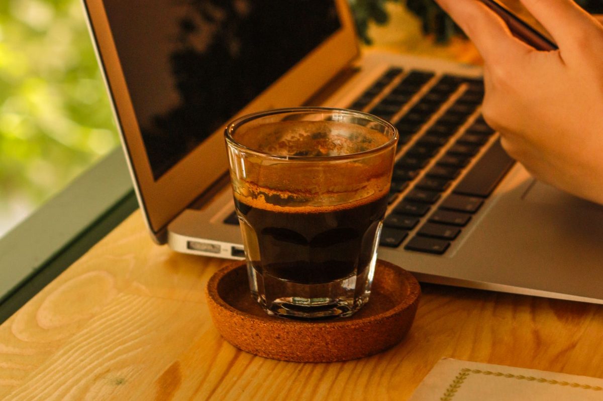 Hand holding a phone beside an open laptop, espresso glass, and a book on a wooden table