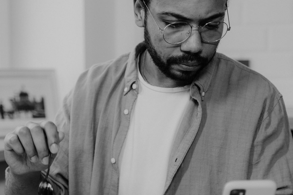 Man with glasses looking at a smartphone while stirring a coffee cup at a table