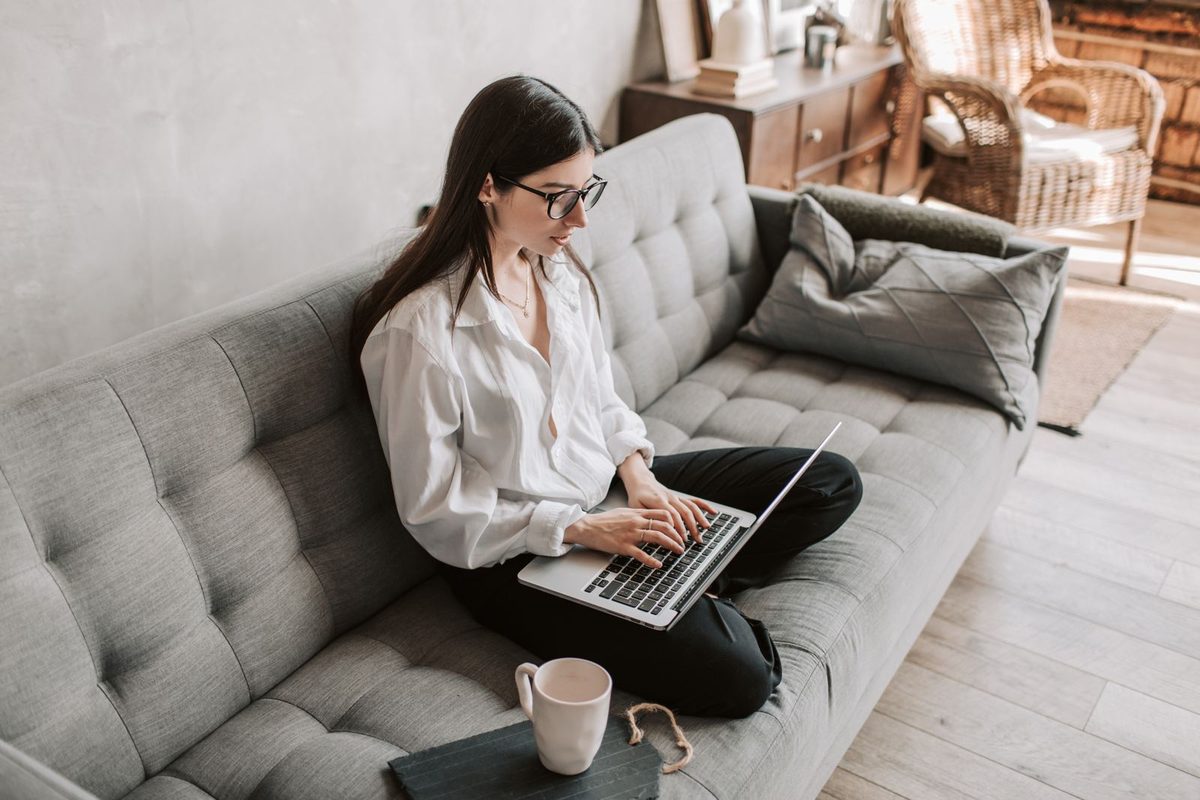 Woman with glasses sitting cross-legged on a sofa typing on a laptop with a mug beside her