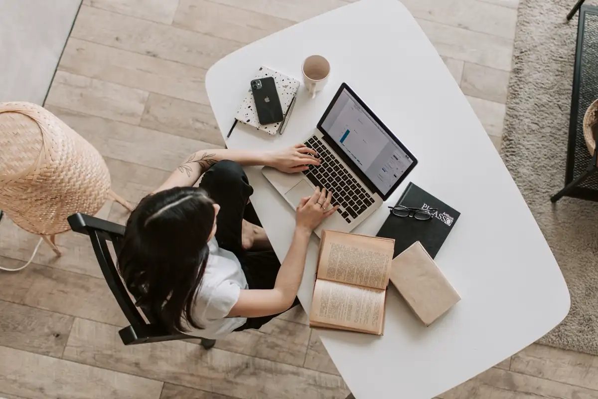 Overhead view of a person at a white desk with a laptop, books, and a smartphone lying face down on a notebook