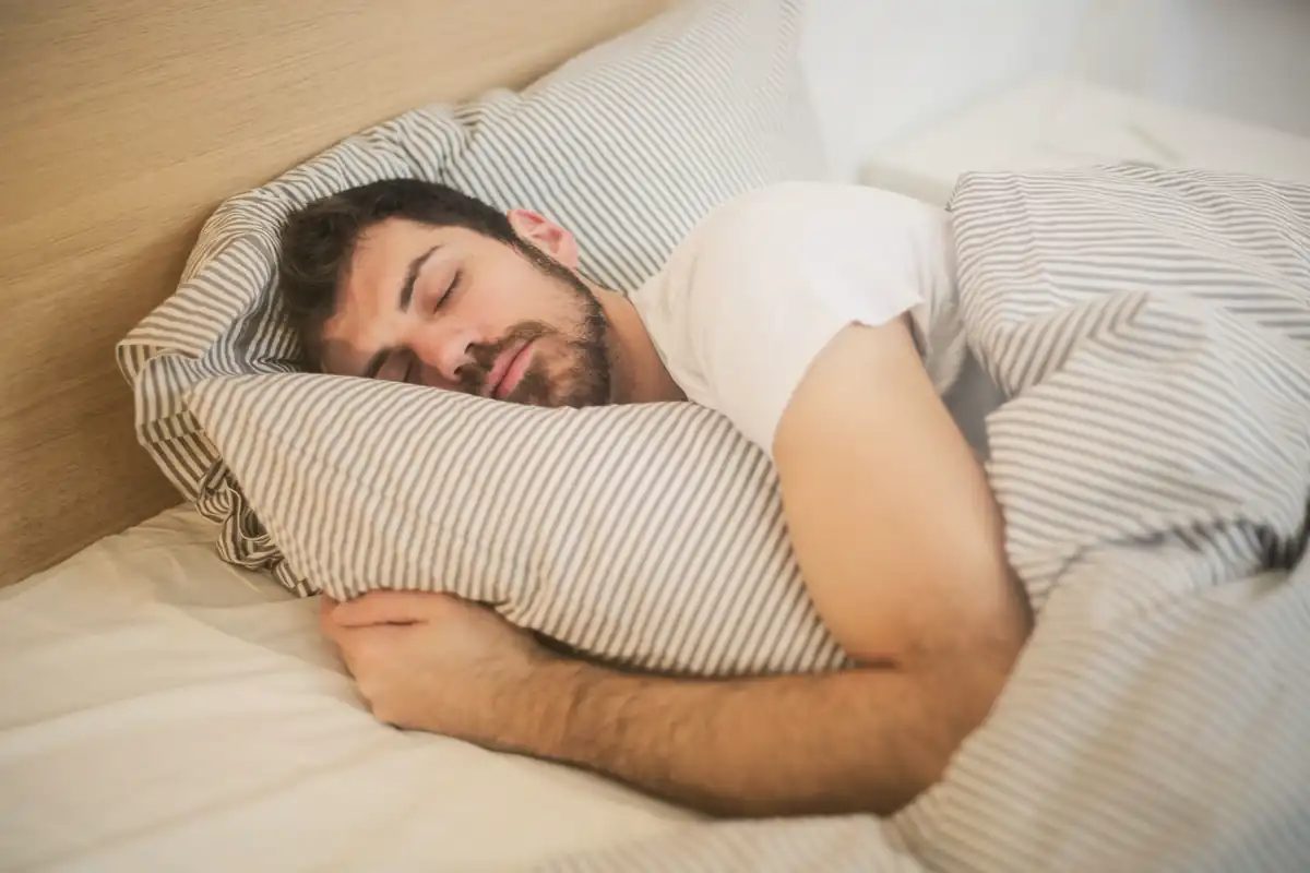 A bearded man sleeping on his side in bed, hugging a pillow under striped bedding
