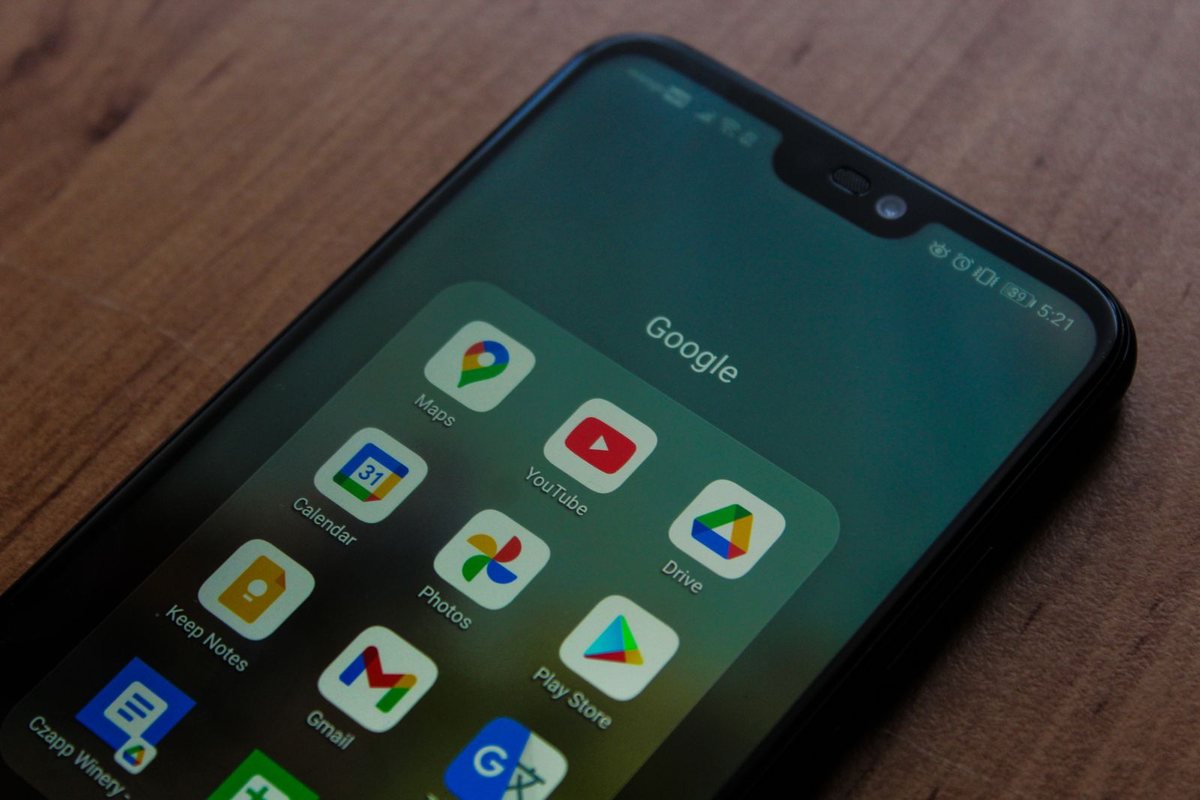 An Android phone on a wooden table, its home screen showing a Google folder with Maps, YouTube, Calendar, Photos, Drive, Keep Notes, Gmail, and Play Store