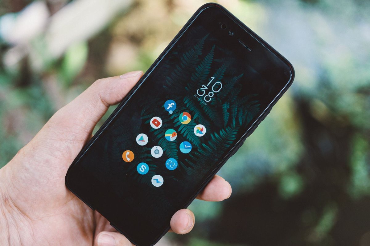 A hand holding an Android phone outdoors, its home screen showing a large clock and a small cluster of round app icons over a dark fern wallpaper