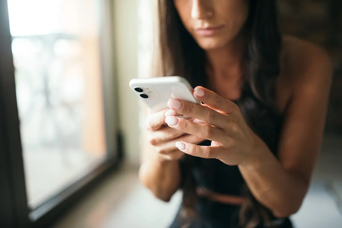A woman sitting by a window, looking down at a phone held in both hands