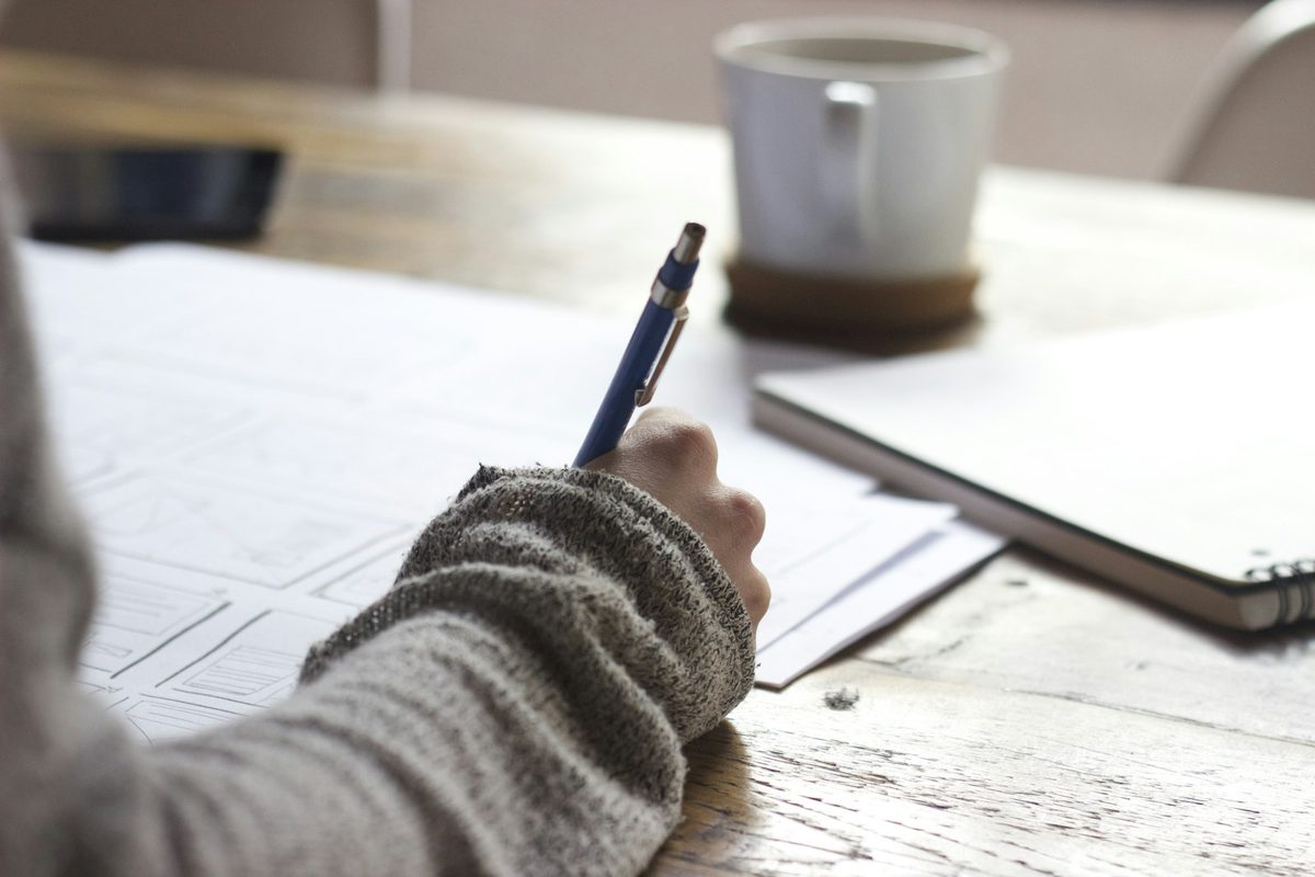 Hand writing on paper with a blue pencil beside a white mug on a wooden desk