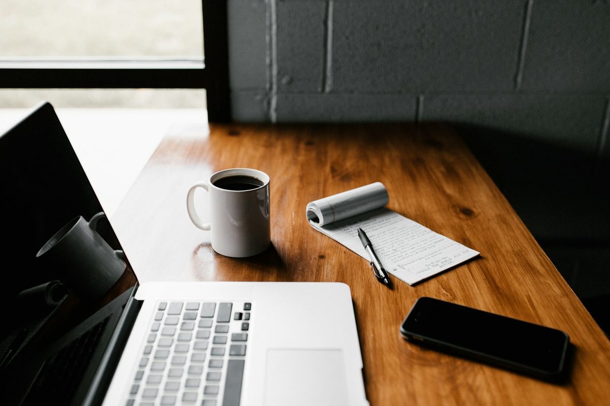 Wooden desk with a laptop keyboard, white coffee mug, notepad with a pen, and a black phone lying face down
