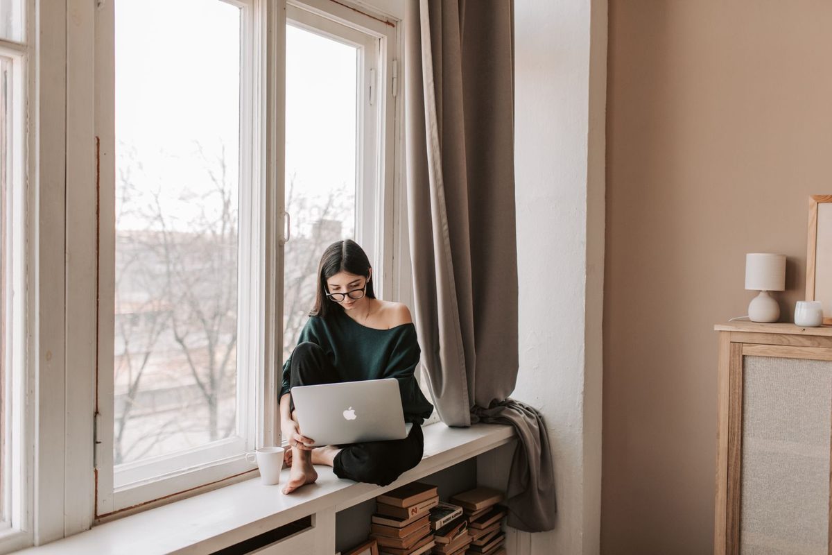 Woman with glasses sitting on a windowsill, focused on a laptop with a mug beside her