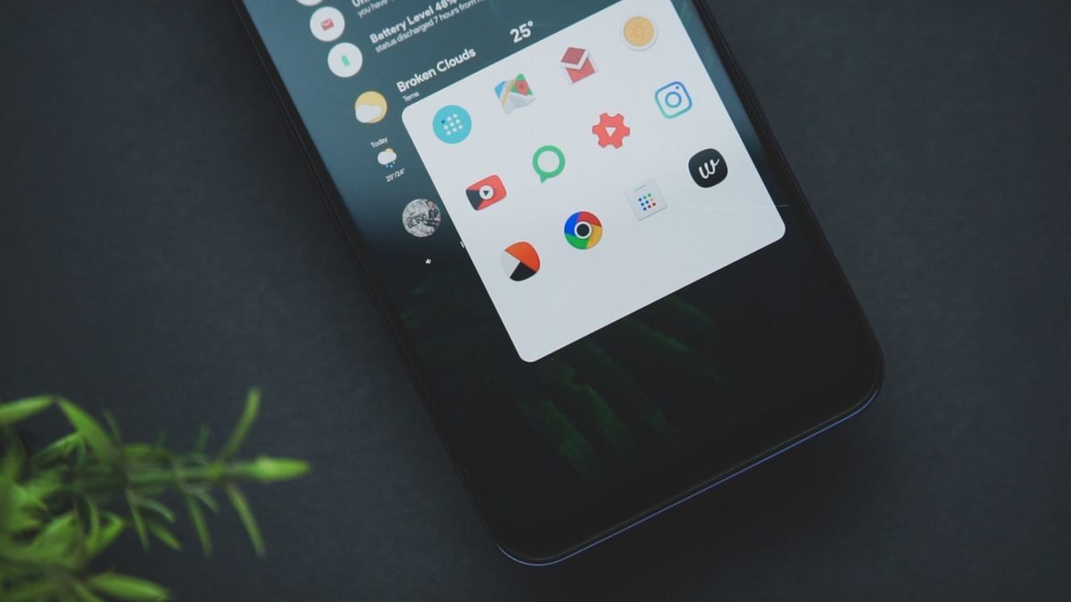 Android phone on a dark table showing app icons on a black background, with a plant in the corner
