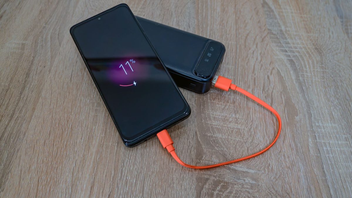 Android phone with a dark charging screen showing 77 percent, plugged into a power bank with an orange cable