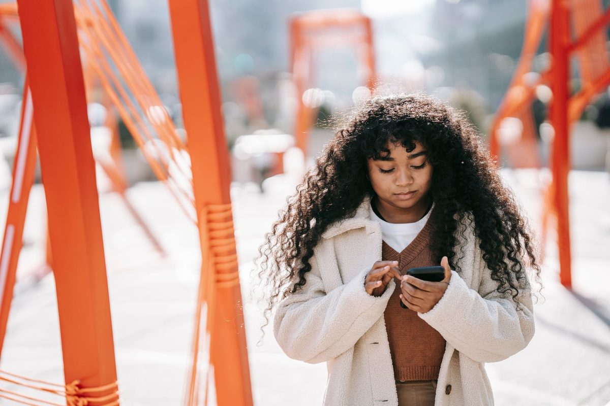 Young woman in a cream coat looking down at her phone on a bright, sunny street