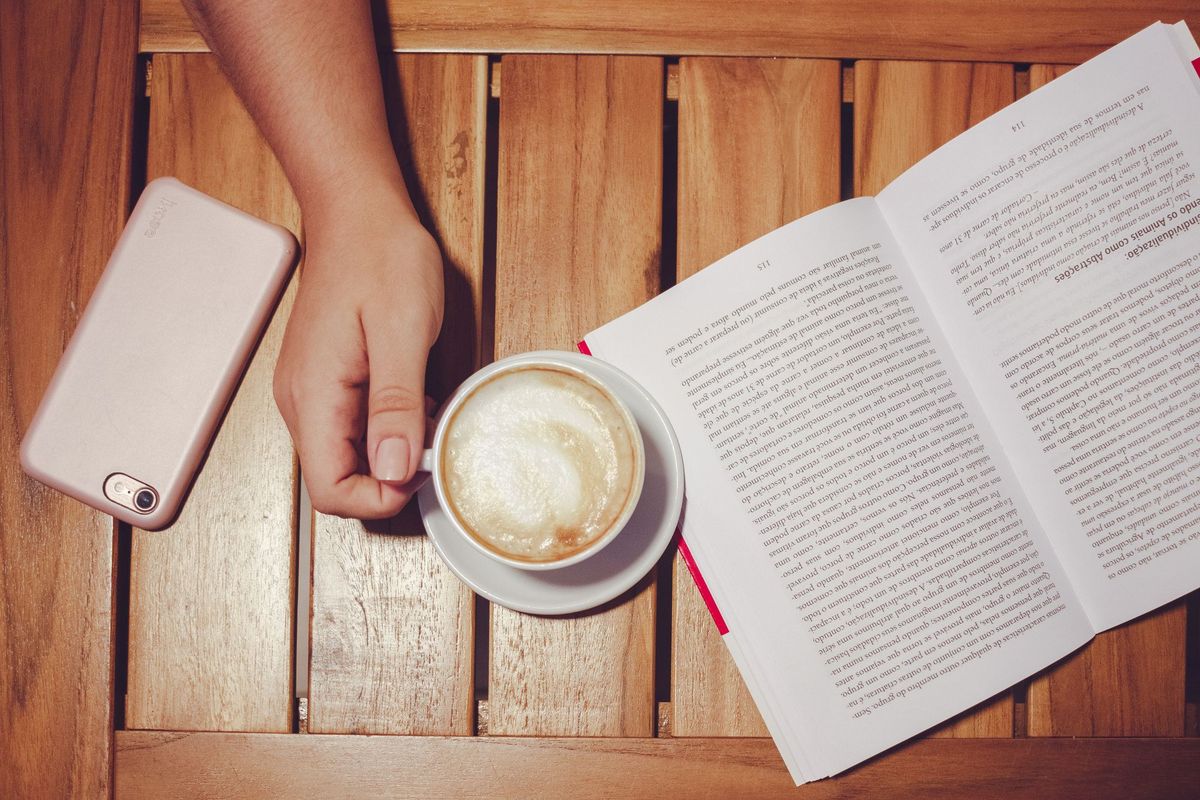 A smartphone lying face down on a wooden table next to an open book and a cup of latte