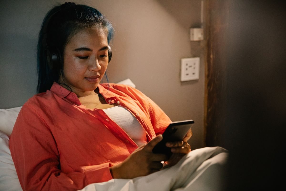 A young woman lying in bed under a blanket, watching her phone with headphones on in a dim room