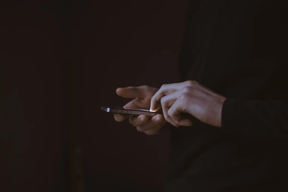 Two hands holding a phone in a dark room, the screen lighting the fingertips