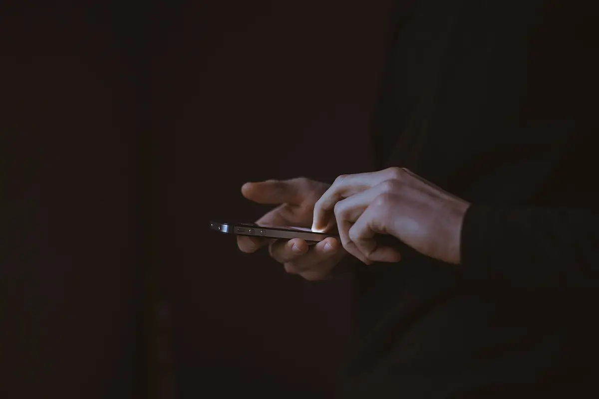 Two hands holding a phone in a dark room, the screen lighting the fingertips