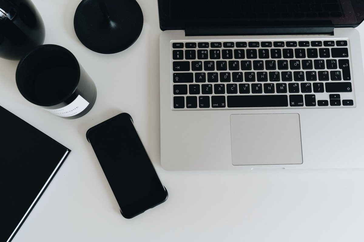 A black smartphone with a dark screen lying next to an open laptop on a white desk, with a mug and a notebook
