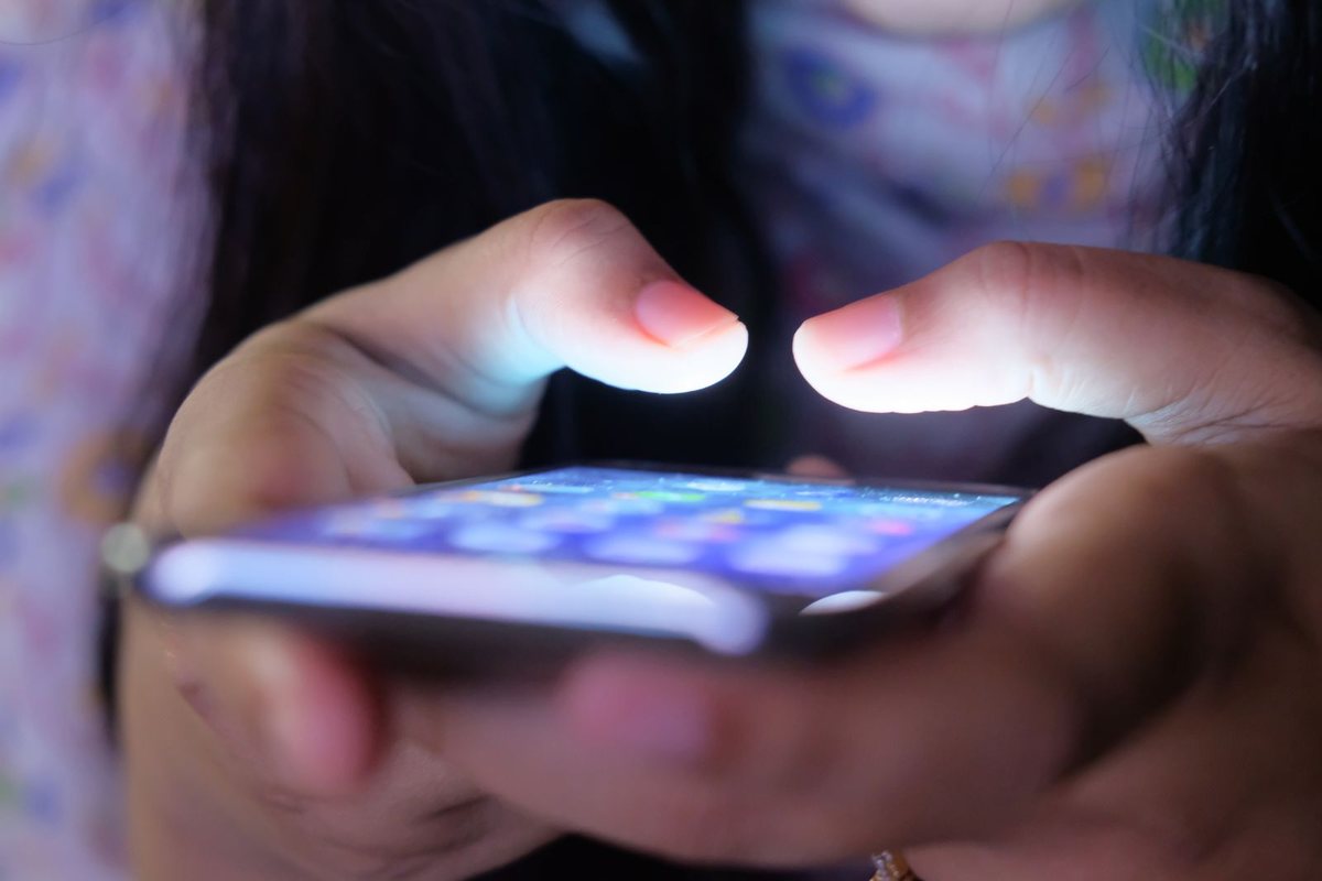 Two hands holding a smartphone in low light, the screen lighting up the fingertips