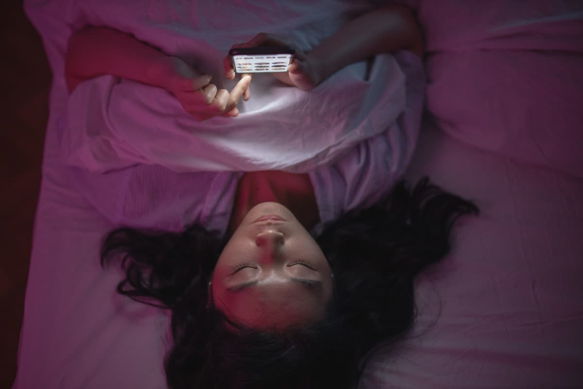 A woman lying in bed in a dim room, scrolling on a smartphone held above her face