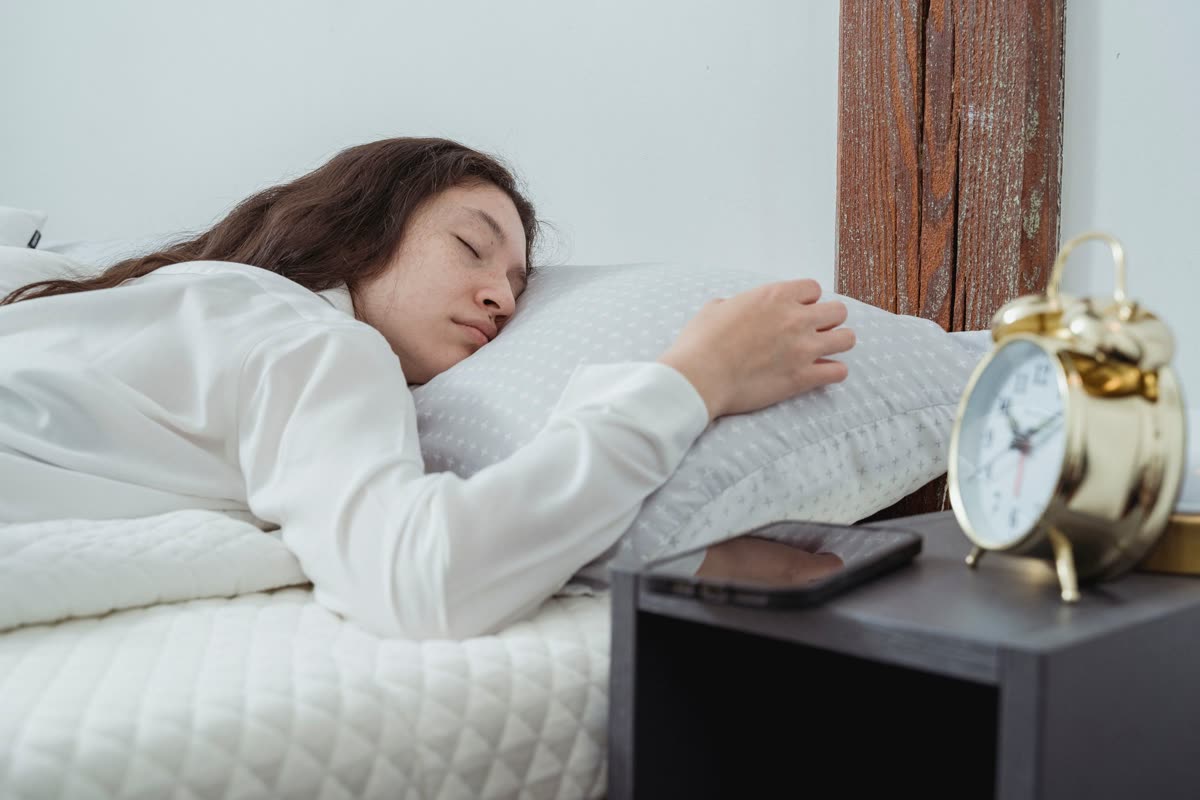A woman asleep in bed with a smartphone lying face down on the bedside table next to an alarm clock