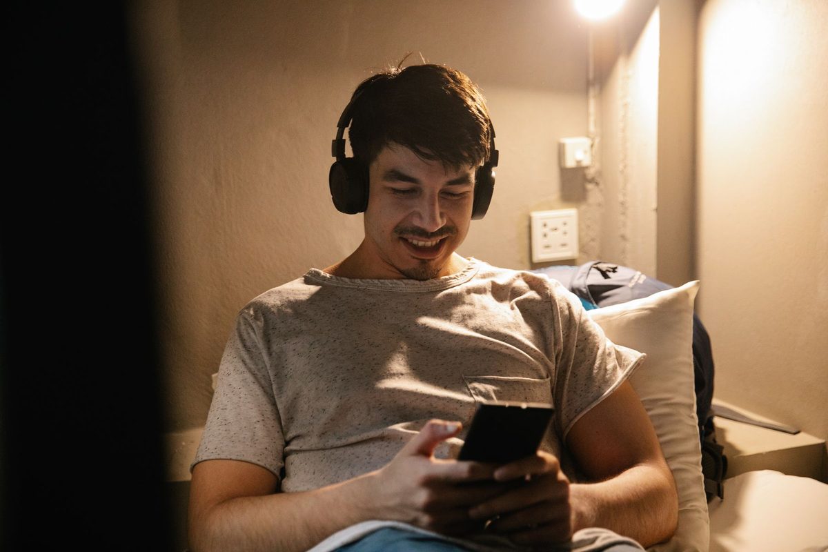 A man in headphones sitting up in bed in a dim room, smiling at his phone